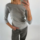 En person tager en selfie i et spejl. De bærer en grå sweater og bukser, står med én hånd i lommen og har et guldarmbånd. Baggrunden er neutral.
