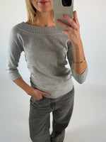 En person tager en selfie i et spejl. De bærer en grå sweater og bukser, står med én hånd i lommen og har et guldarmbånd. Baggrunden er neutral.