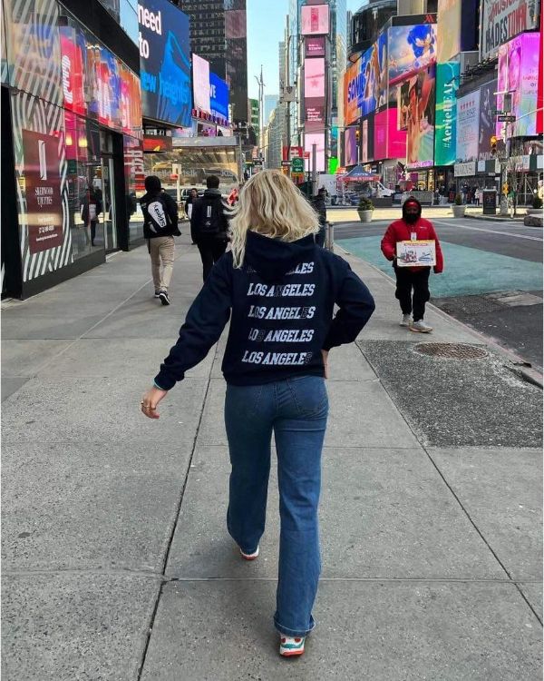 En person med en "Los Angeles"-trøje går på en travl byfortov om dagen. Omkring ses andre fodgængere, skyskrabere og store reklameskærme.