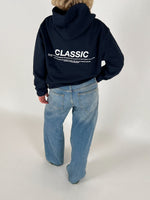 En person iført en mørk hættetrøje med teksten "CLASSIC" og blå jeans står i et lyst rum. Teksten på hættetrøjen fortsætter med mindre skrift under hovedordet.