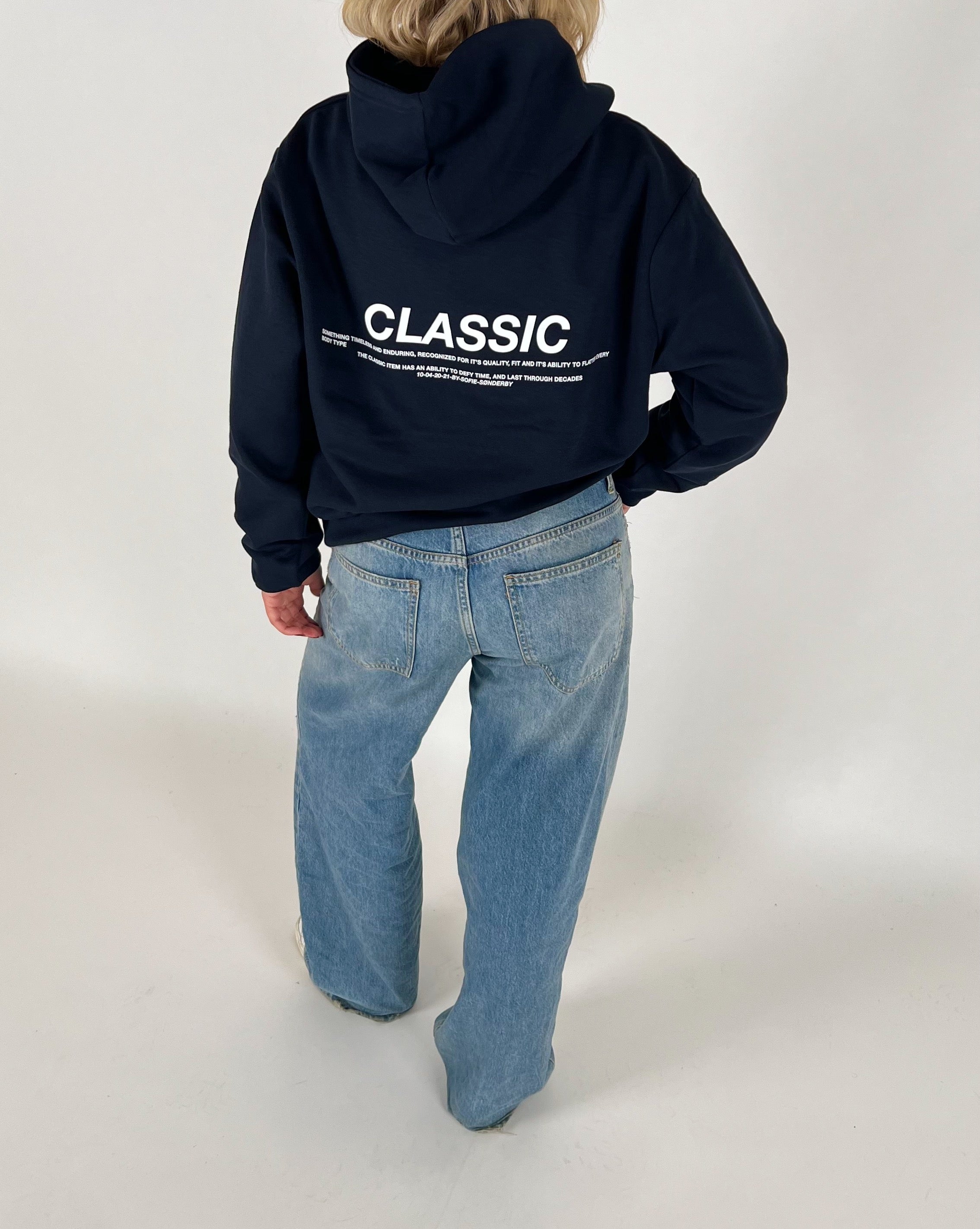 En person iført en mørk hættetrøje med teksten "CLASSIC" og blå jeans står i et lyst rum. Teksten på hættetrøjen fortsætter med mindre skrift under hovedordet.