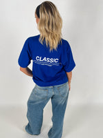 En person står med ryggen til iført en blå T-shirt, hvor der står "CLASSIC" i hvid tekst og løse jeans, mod en neutral baggrund.