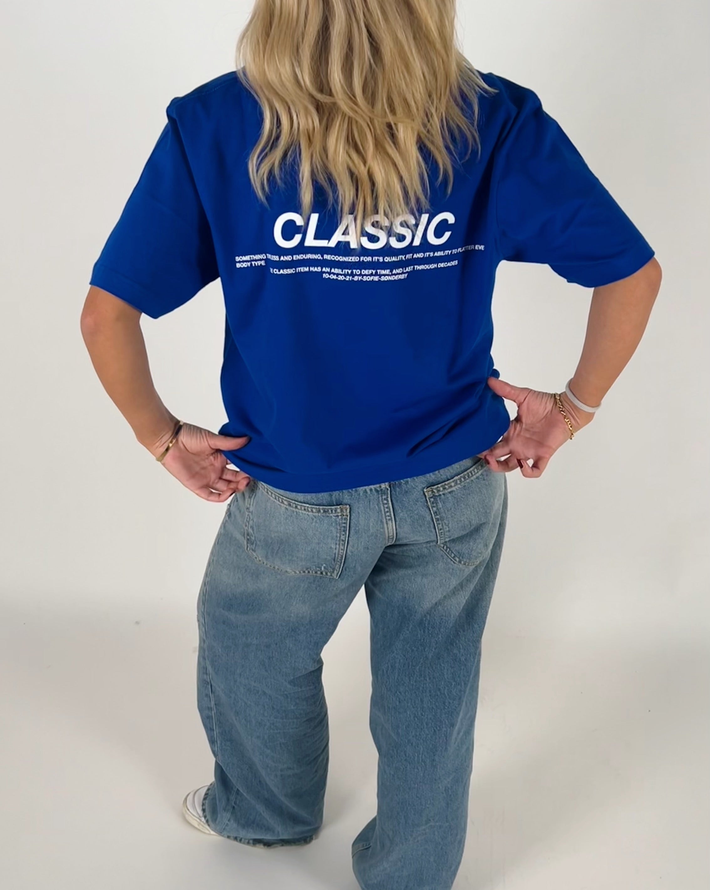 En person iført en blå T-shirt med teksten "CLASSIC" og en længere tekst nedenunder står med hænderne på hofterne i et lyst rum. De har også lyse jeans på.