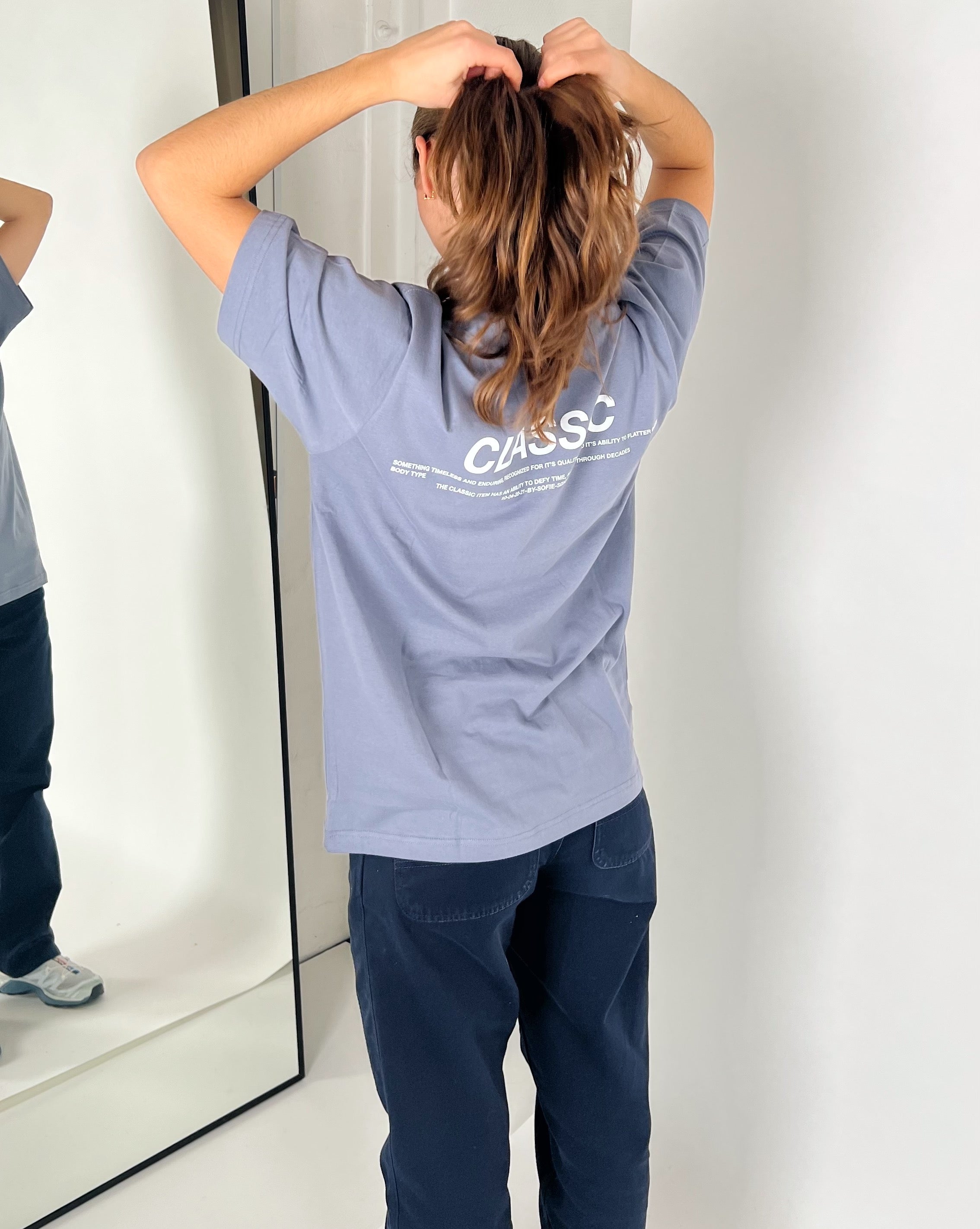 En person i en stone t-shirt arrangerer håret foran et spejl. Spejlingen viser også teksten "CLASSIC" og anden mindre tekst på t-shirtens ryg.