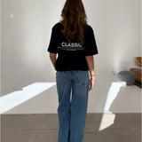 En person med langt hår og blå jeans står stille med ryggen til, iført en sort T-shirt med teksten "CLASSIC" i et minimalistisk, lyst rum.
