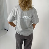 En person iført en grå T-shirt og mørke bukser står med ryggen til. På T-shirten står teksten "CLASSIC". Baggrunden er lys og enkel.