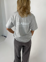 En person iført en grå T-shirt og mørke bukser står med ryggen til. På T-shirten står teksten "CLASSIC". Baggrunden er lys og enkel.