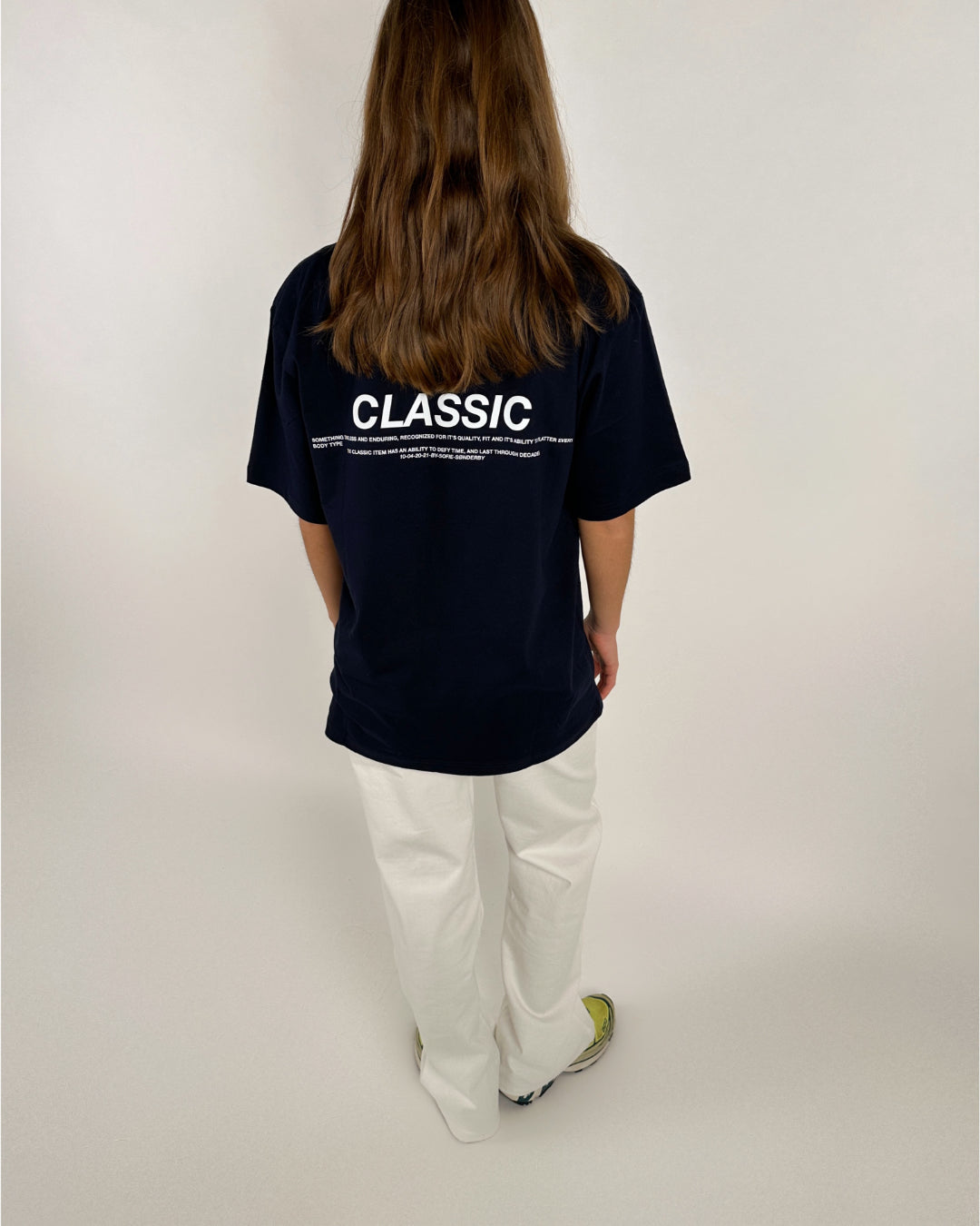 Person bærer en sort T-shirt med teksten "CLASSIC" i hvid, samt flere små skrifttyper nedenunder. Personen står med ryggen til iført hvide bukser og farverige sko i et neutralt rum.