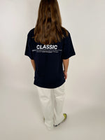 Person bærer en sort T-shirt med teksten "CLASSIC" i hvid, samt flere små skrifttyper nedenunder. Personen står med ryggen til iført hvide bukser og farverige sko i et neutralt rum.