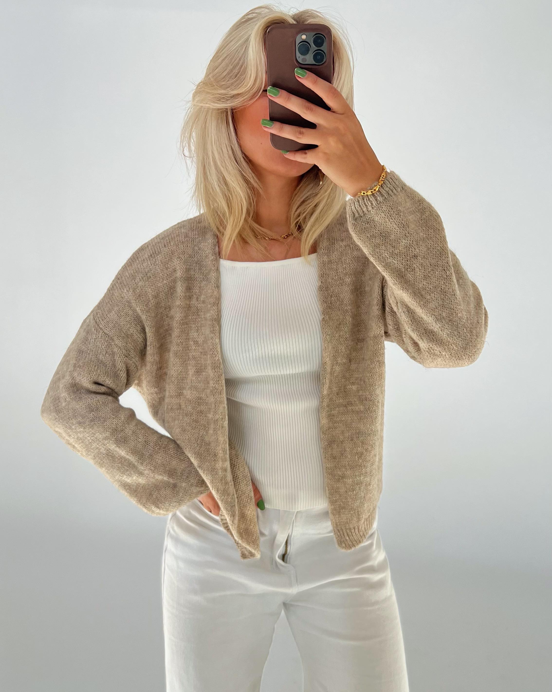Person holder en telefon op foran ansigtet, iført en beige cardigan og hvide bukser, står i et neutralt oplyst rum. Personen har grønmalede negle.