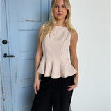 En kvinde står foran en lys blå dør iført en lys beige top og sorte bukser, med en neutral baggrund.