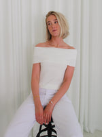 Person sidder på en sort stol iført en hvid, off-shoulder top og hvide bukser, med hænderne hvilende i skødet. Baggrunden er hvide gardiner.