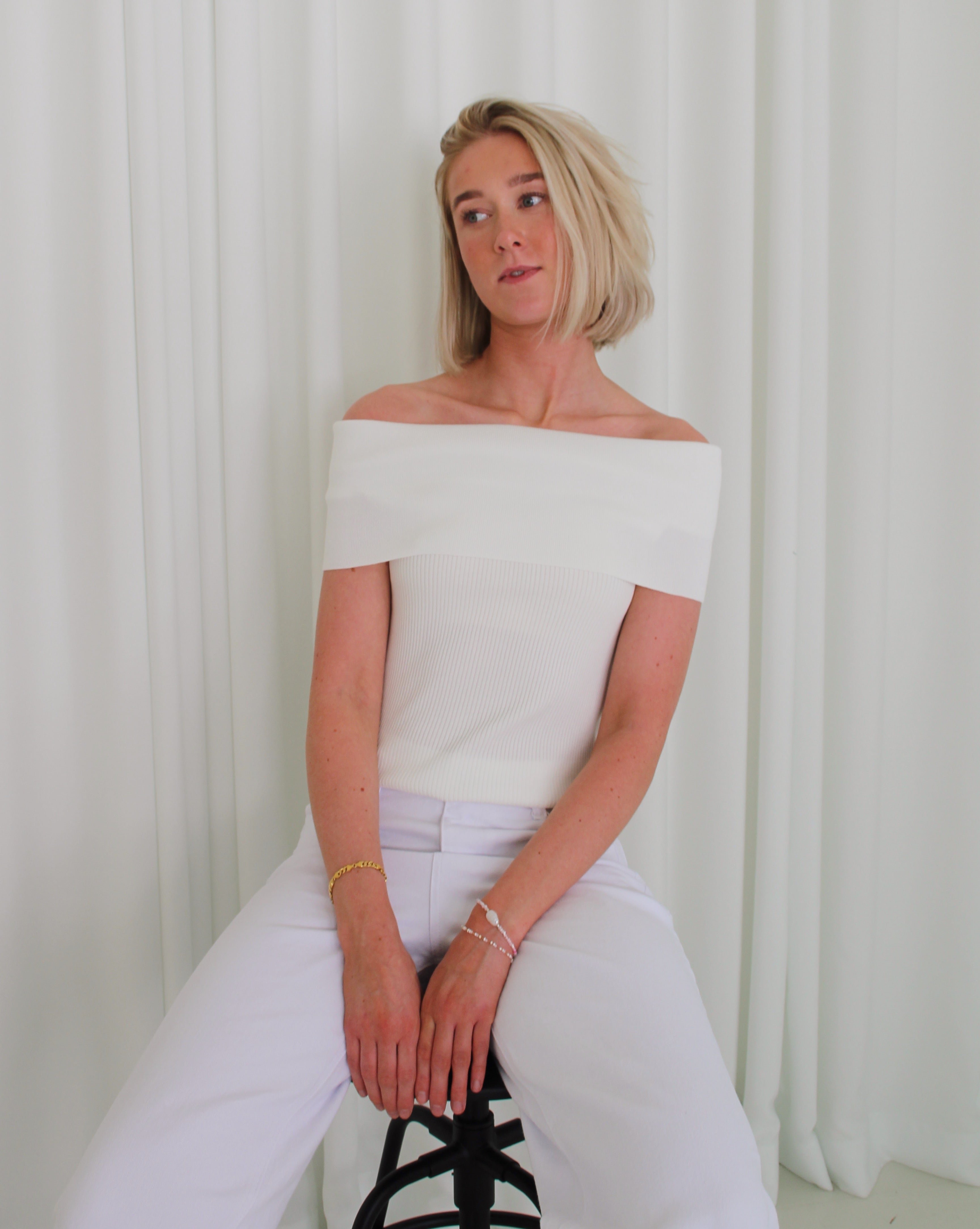 Person sidder på en sort stol iført en hvid, off-shoulder top og hvide bukser, med hænderne hvilende i skødet. Baggrunden er hvide gardiner.