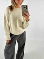 Person i cremefarvet sweater tager et billede med en telefon. De har mørke bukser på og poserer i et neutralt rum.