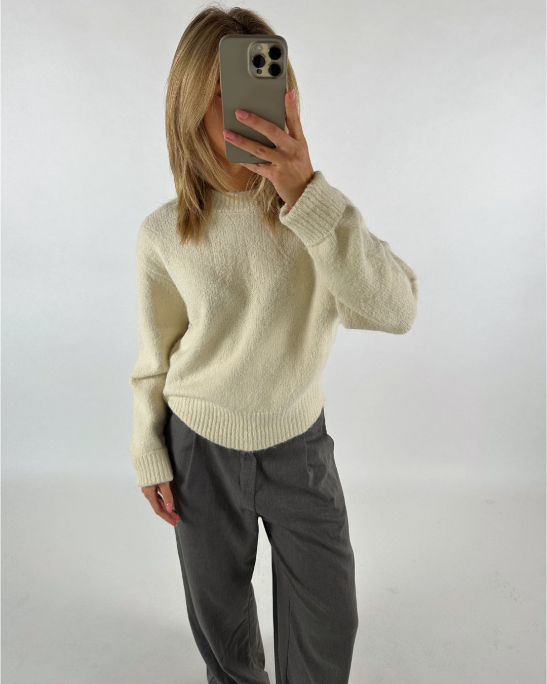 Person tager et spejlbillede, iført en cremefarvet sweater og grå bukser, i et hvidt rum. Personen holder en mobiltelefon op til ansigtet og har skulderlangt hår.