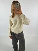 Person tager et spejlbillede, iført en cremefarvet sweater og grå bukser, i et hvidt rum. Personen holder en mobiltelefon op til ansigtet og har skulderlangt hår.