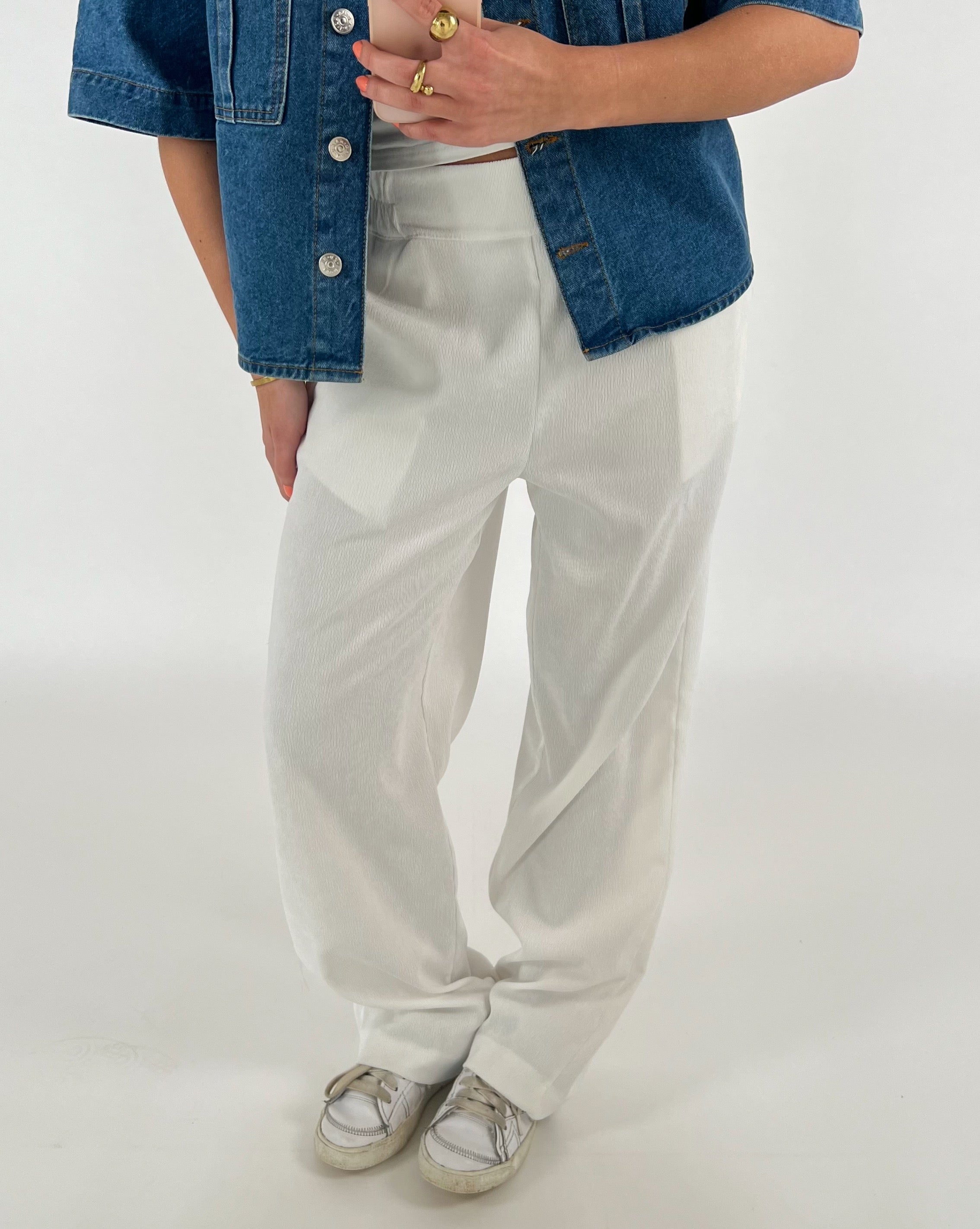 En person iført afslappede, hvide bukser og en blå denimjakke står i en minimalistisk, lys baggrund, mens vedkommende holder en smartphone i hånden.