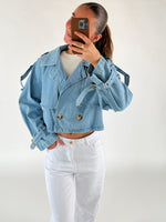 En person iført en lyseblå denimjakke tager en selfie med sin telefon foran et hvidt baggrund. Jakken har store knapper og bukserne er hvide.