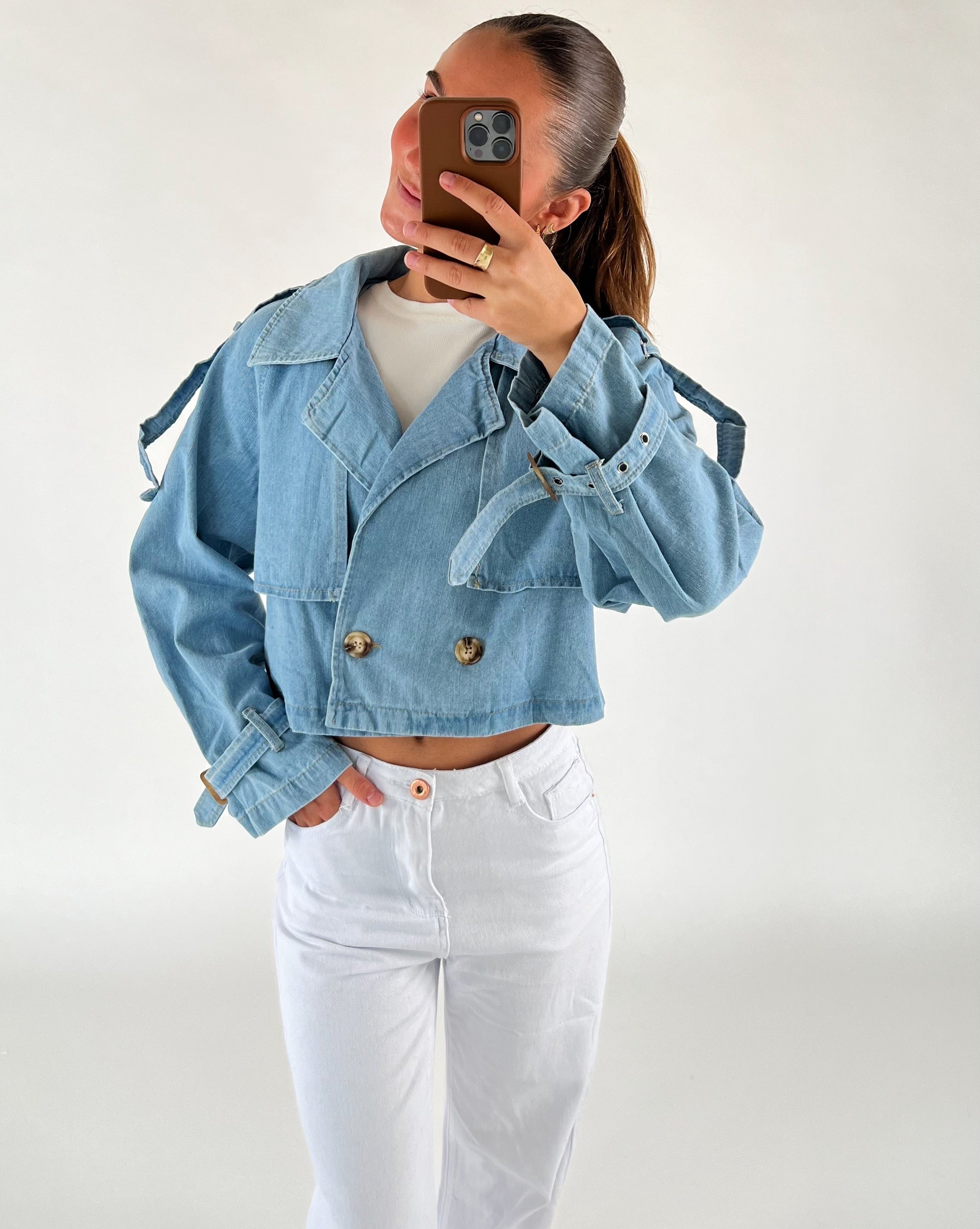En person iført en lyseblå denimjakke tager en selfie med sin telefon foran et hvidt baggrund. Jakken har store knapper og bukserne er hvide.