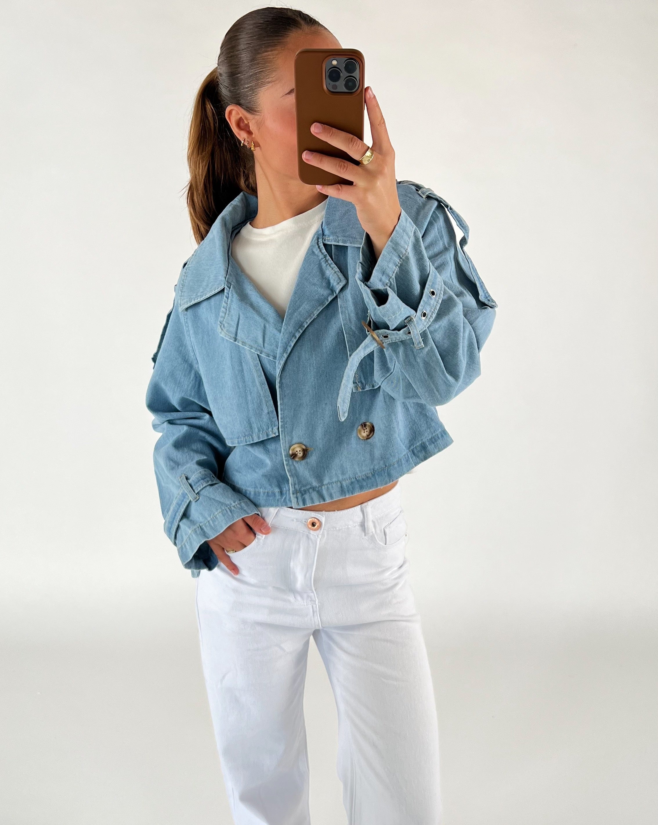 En person iført en lyseblå denimjakke og hvide bukser tager et spejlbillede med en telefon i brun cover i en lys baggrund.