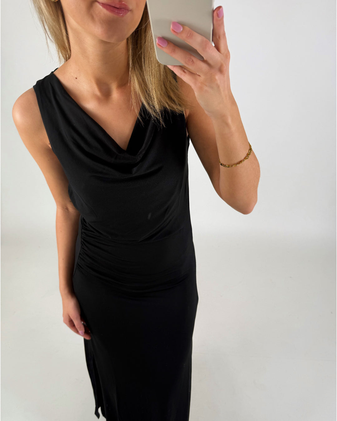 Kvinde i sort kjole tager selfie med mobiltelefon i et lyst rum. Hun har lyse hår, pink negle og et guldfarvet armbånd.