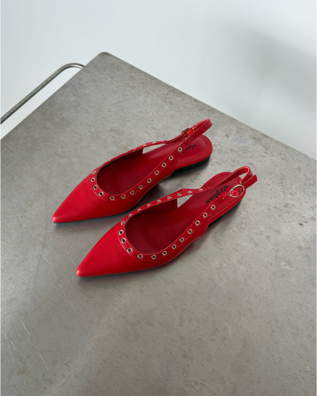 Røde slingback-sko med nitter ligger stille på en grå bordflade. Indersålen har teksten "sezane."