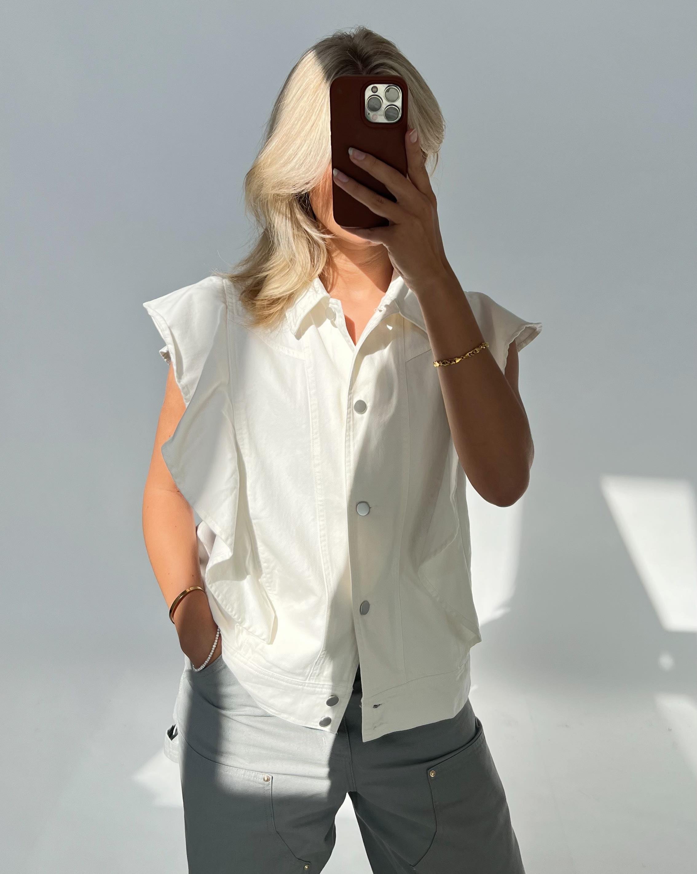 Person tager en selfie med mobiltelefon, iført hvid ærmeløs skjorte og lysegrå bukser, i et lyst rum med bløde skygger.