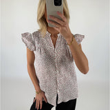Kvinde iført blomstret bluse tager selfie med mobiltelefon i et lyst rum. Hun har sort tøj på og bærer et guldarmbånd.