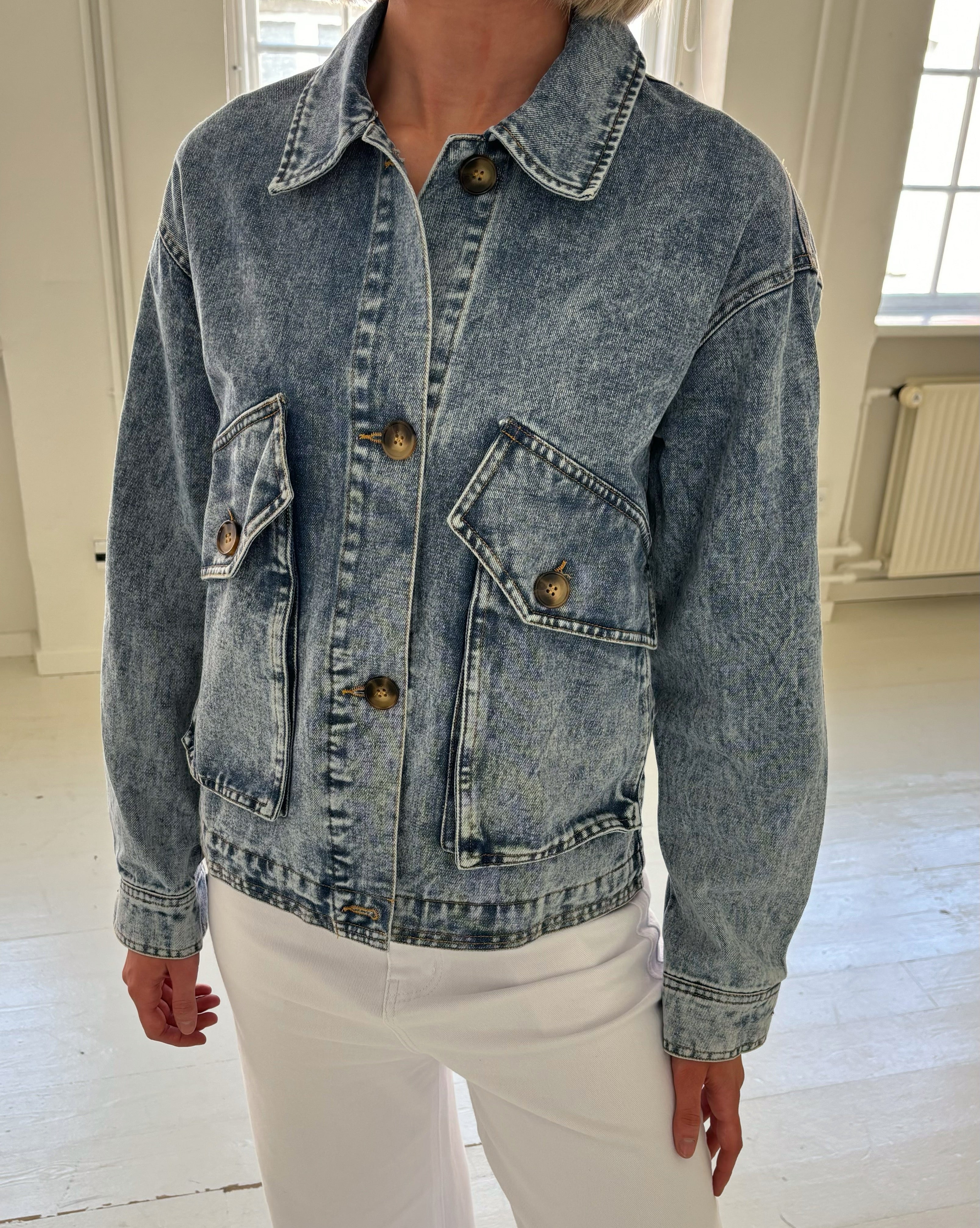 En person bærer en blå denimjakke med brune knapper og store lommer. Personen står i et lyst rum med hvidmalede gulve og vægge.