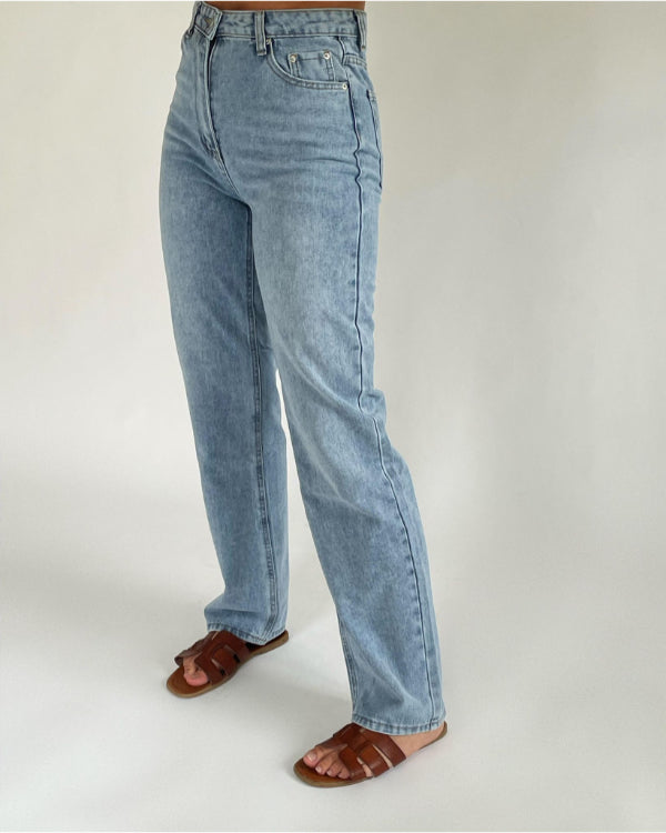Lyseblå jeans bæres af en person, stående oprejst, iført brune sandaler. Baggrunden er enkel og ensfarvet, hvilket fremhæver tøjets detaljer.