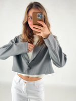 En person iført en grå cropped jakke tager en selfie med en smartphone. Personen har hvidt tøj på under jakken og ringen på venstre hånd. Baggrunden er neutral.