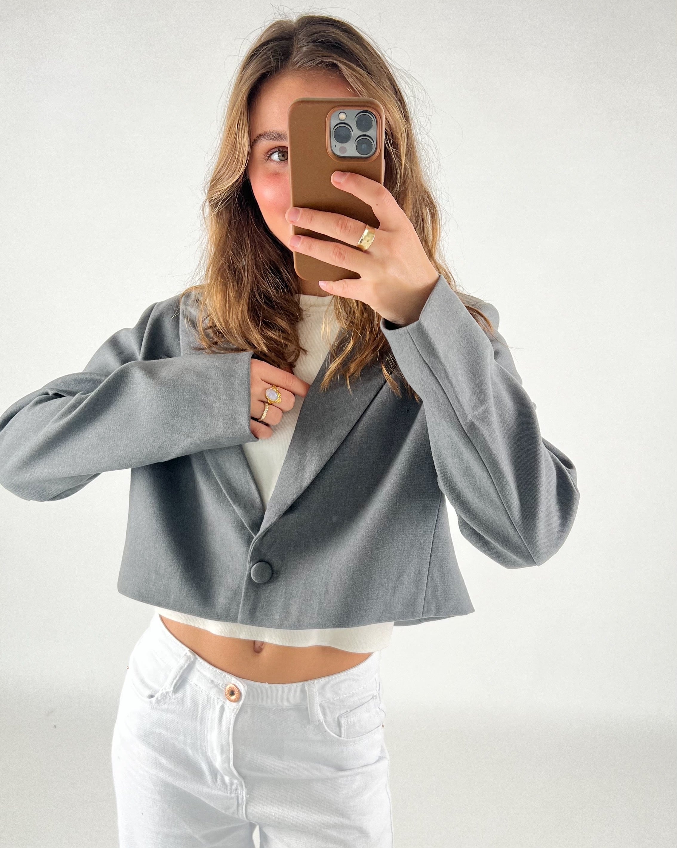 En person iført en grå cropped jakke tager en selfie med en smartphone. Personen har hvidt tøj på under jakken og ringen på venstre hånd. Baggrunden er neutral.