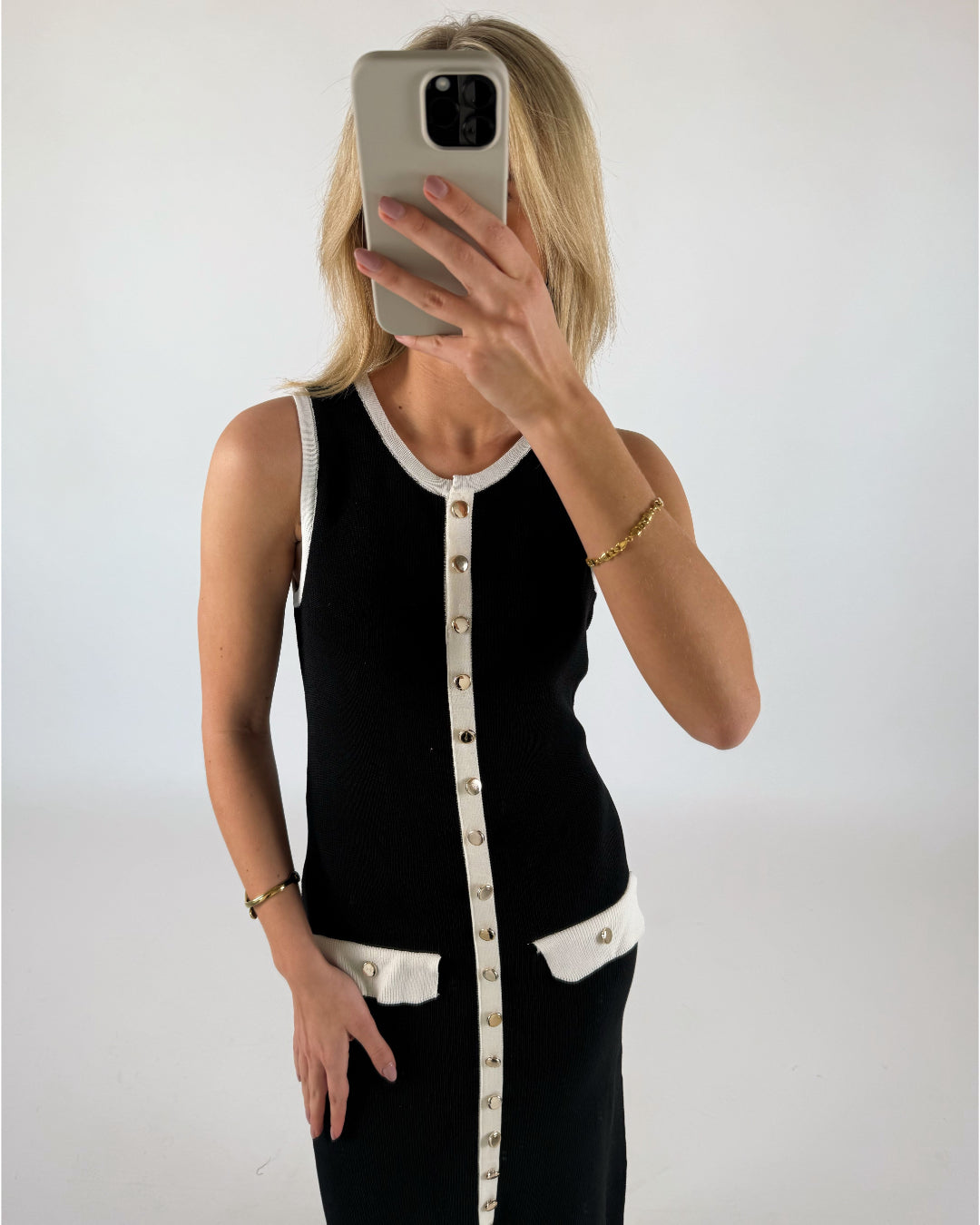 Person tager selfie med mobiltelefon, bærer sort kjole med hvide kanter og guldknapper, står i et lyst rum.