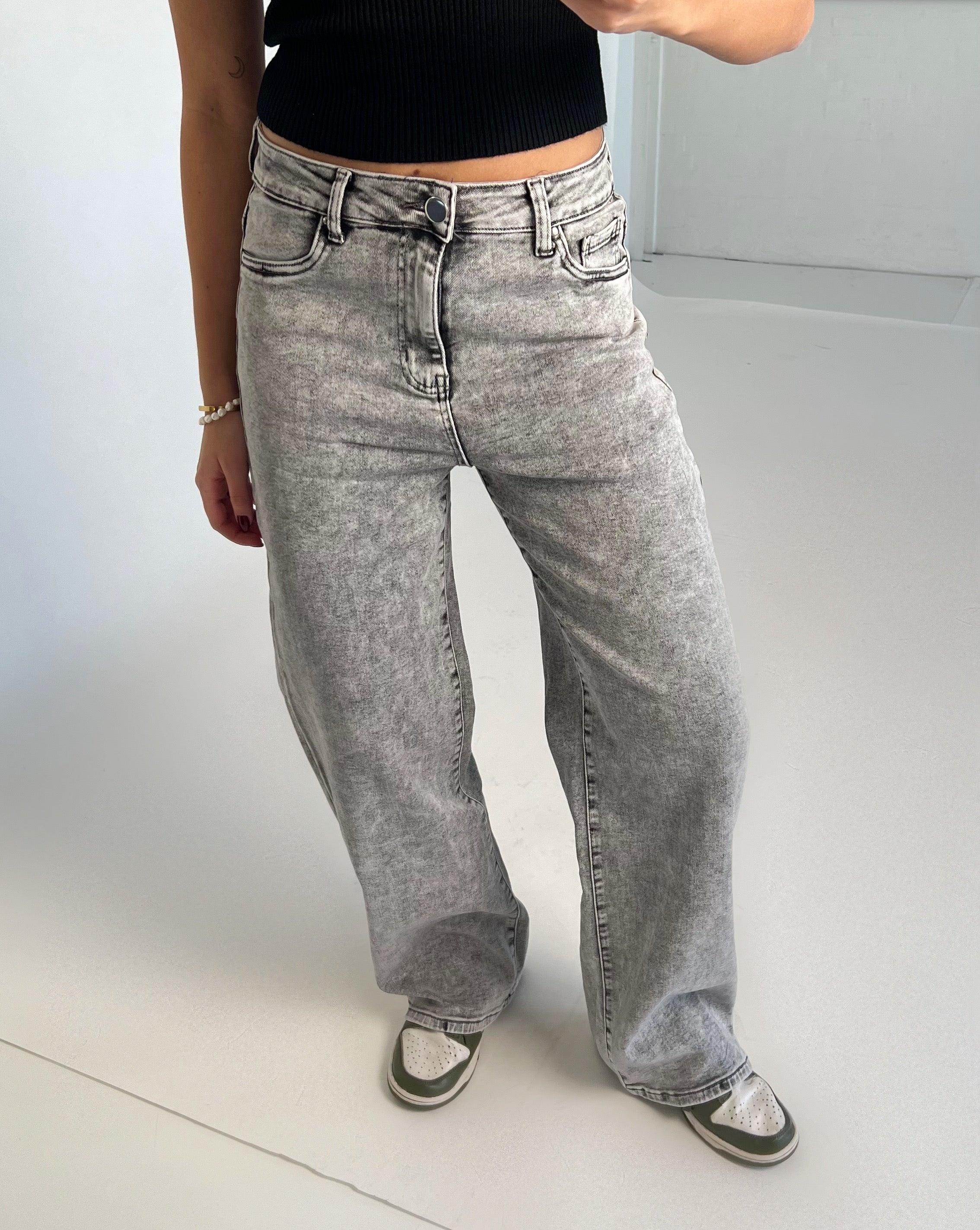En person iført lysegrå, løse cowboystofbukser står oprejst på et lysfarvet gulv. Personen har en sort top på og bærer sneakers. Hintergrund er minimalistisk og lys.