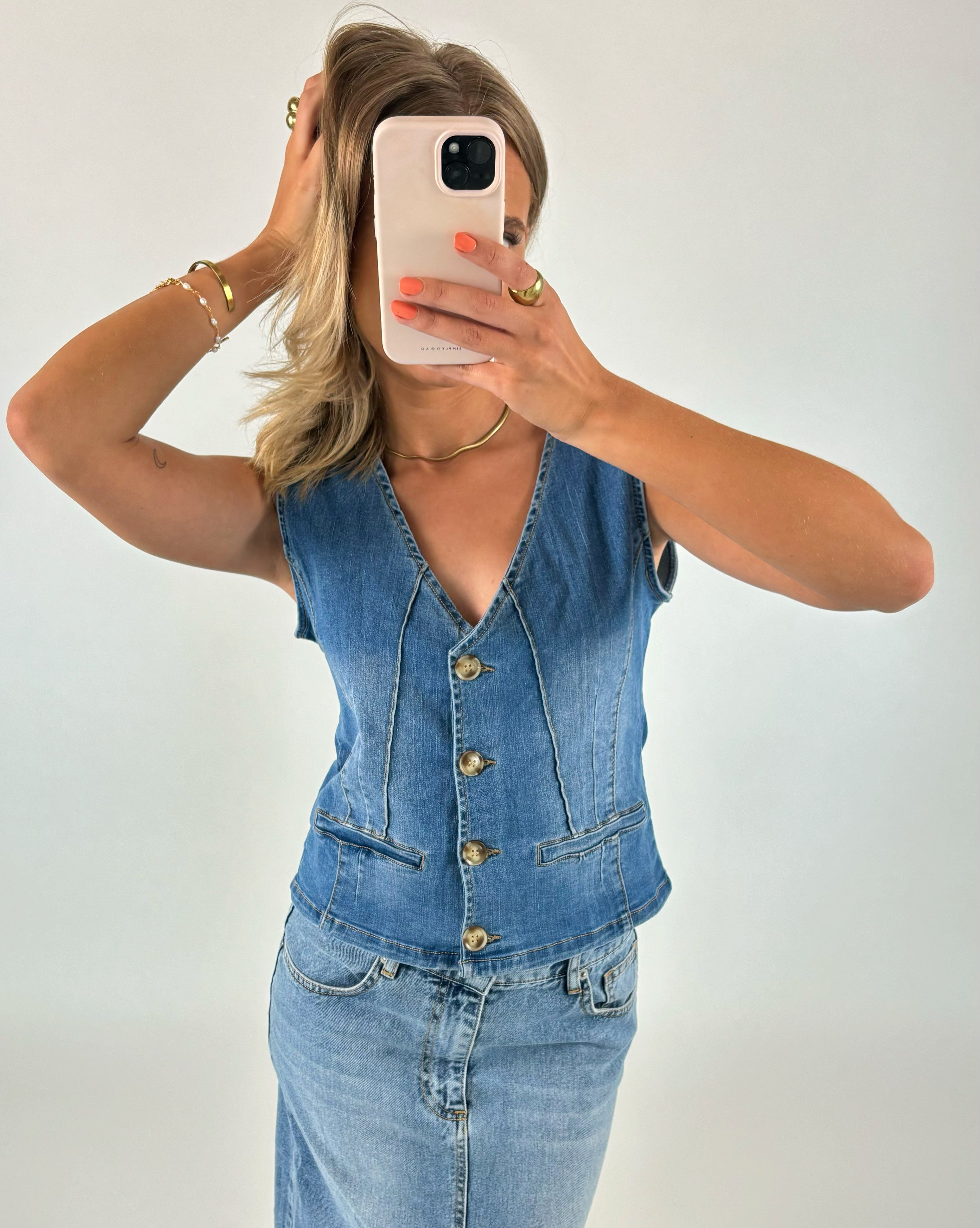 En person iført denimvest og nederdel tager et spejlbillede med en mobiltelefon. Personen har hånden i håret og bærer sølvsmykker. Baggrunden er neutral.