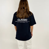 Person i sort T-shirt står med ryggen til, iført hvide bukser. T-shirten har teksten: "CLASSIC" efterfulgt af mindre tekst. Baggrunden er lys og enkel.