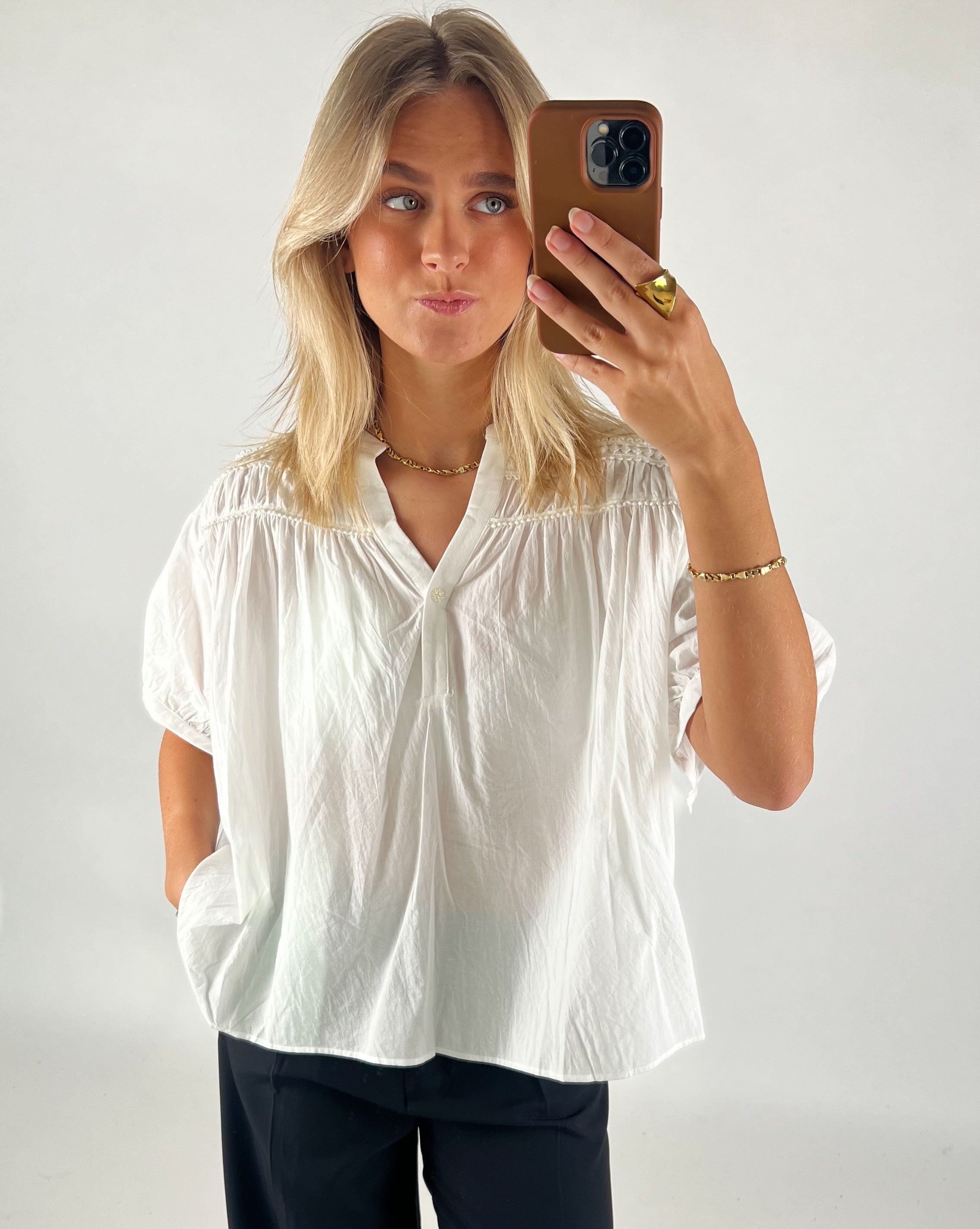 Person tager en selfie med en smartphone. De bærer en hvid bluse og mørke bukser, står foran en neutral, lys baggrund. Personen har lyst hår og bærer guldsmykker.