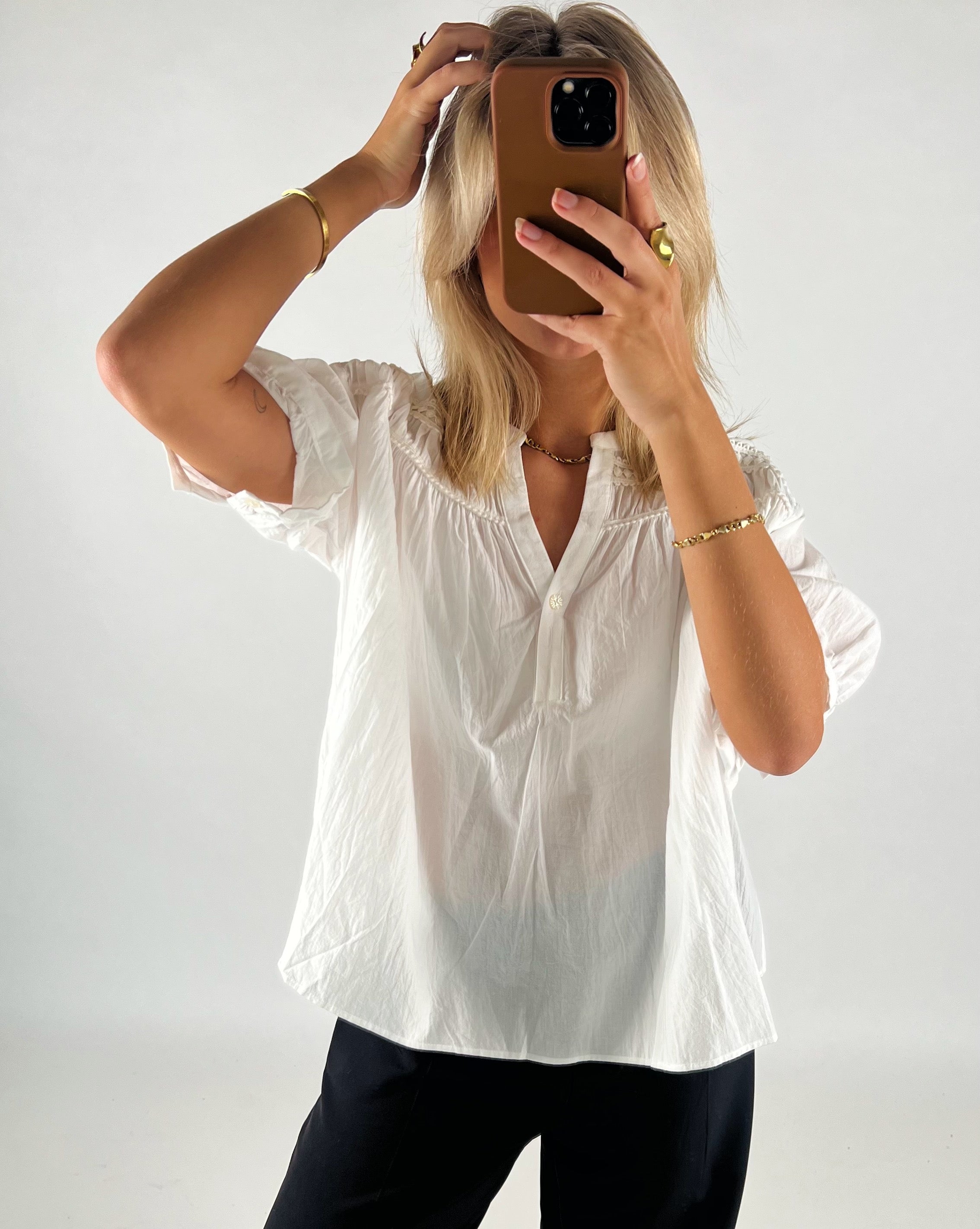 En person tager en selfie med en mobiltelefon, mens de står i et lyst rum. Personen bærer en hvid skjorte og sort bukser, med nogle smykker på håndleddet.