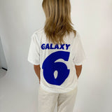 En person med skulderlangt hår står med ryggen til, iført en hvid t-shirt med blå tekst "GALAXY 6" i en lys baggrund.