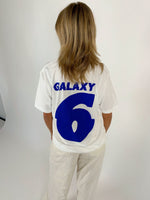 En person med skulderlangt hår står med ryggen til, iført en hvid t-shirt med blå tekst "GALAXY 6" i en lys baggrund.