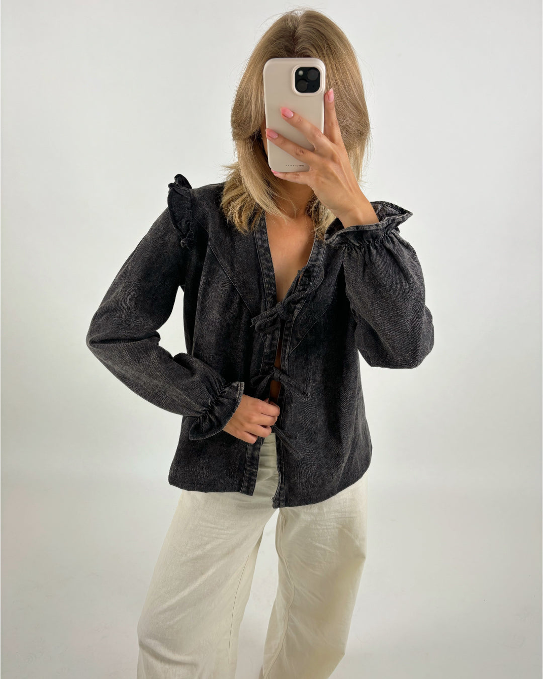 En person tager en selfie med en smartphone, iført en mørk bluse og lyse bukser i et neutralt belyst rum.