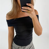 Person tager et spejlbillede iført sort off-shoulder top og grå jeans. De holder en telefon foran ansigtet i en lys, minimalistisk baggrund.