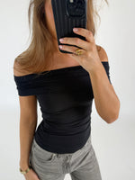 Person tager et spejlbillede iført sort off-shoulder top og grå jeans. De holder en telefon foran ansigtet i en lys, minimalistisk baggrund.