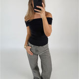 En person tager et spejlbillede, iført en sort, skulderfri top og grå jeans, mens de står i et minimalistisk, lyst rum med en neutral væg.