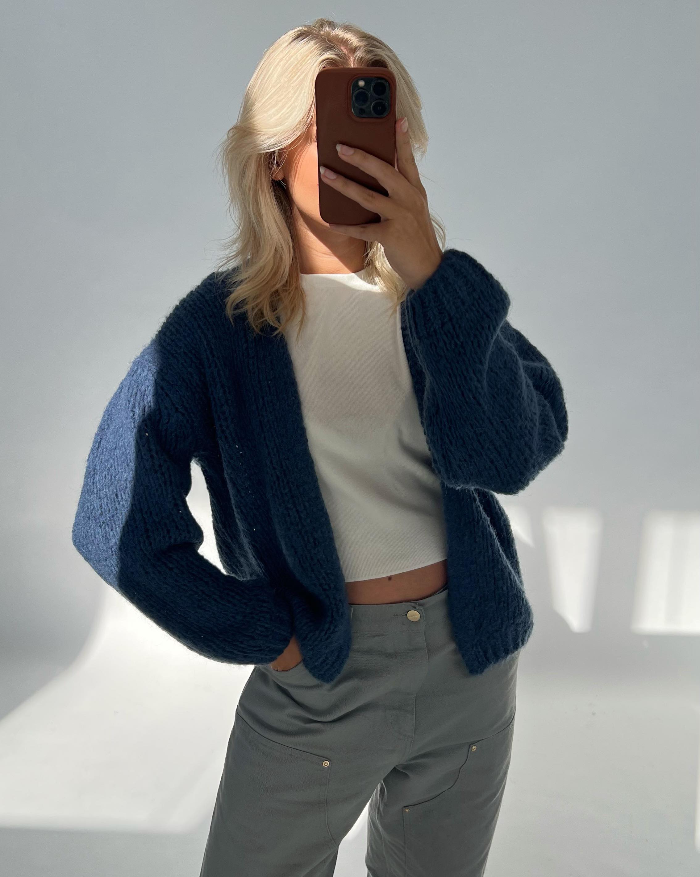 Person tager et spejlselfie iført en hvid top, mørkeblå striktrøje og grå bukser. Baggrunden er neutral, med blødt lys der skaber skygger på væggen.