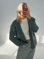 Person tager et billede med mobiltelefon, iført en striktrøje over en hvid T-shirt, stående i et rum med gråt sollys.