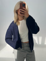 En person tager et spejlbillede og holder en mobiltelefon foran ansigtet. Personen har lyst hår, en mørk cardigan, en hvid trøje og grå bukser i et lyst rum.