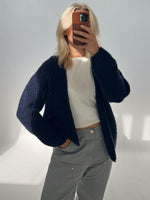 Person tager et spejlbillede med en smartphone, iført en mørk sweater og lyse bukser, i et minimalistisk, lyst rum uden tekst.