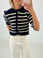 Kvinde iført sort-hvid-stribet cardigan med knapper, fotograferer sig selv med en smartphone. Hun har lyse jeans på og står i et neutralt, lyst rum.