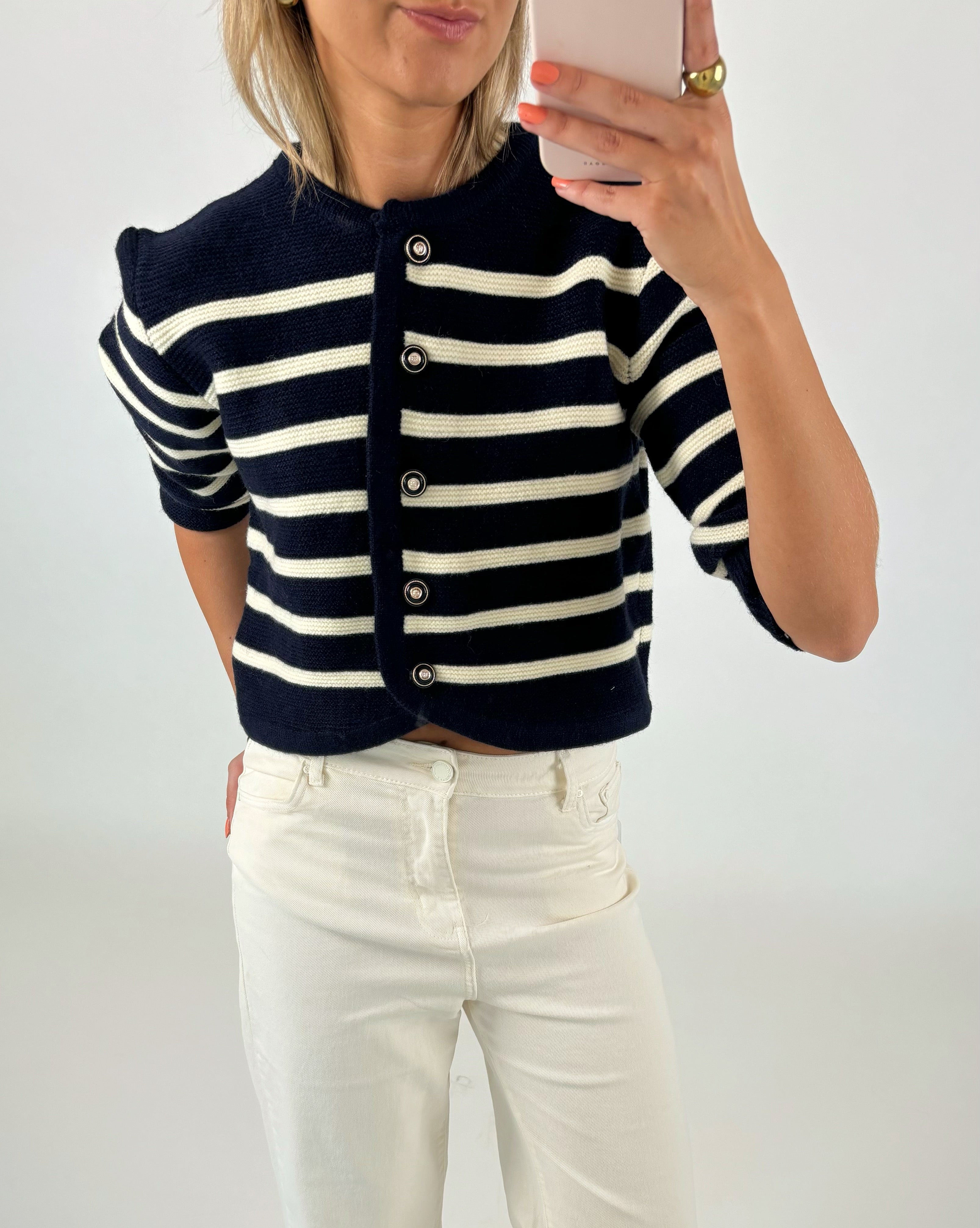 Kvinde iført sort-hvid-stribet cardigan med knapper, fotograferer sig selv med en smartphone. Hun har lyse jeans på og står i et neutralt, lyst rum.